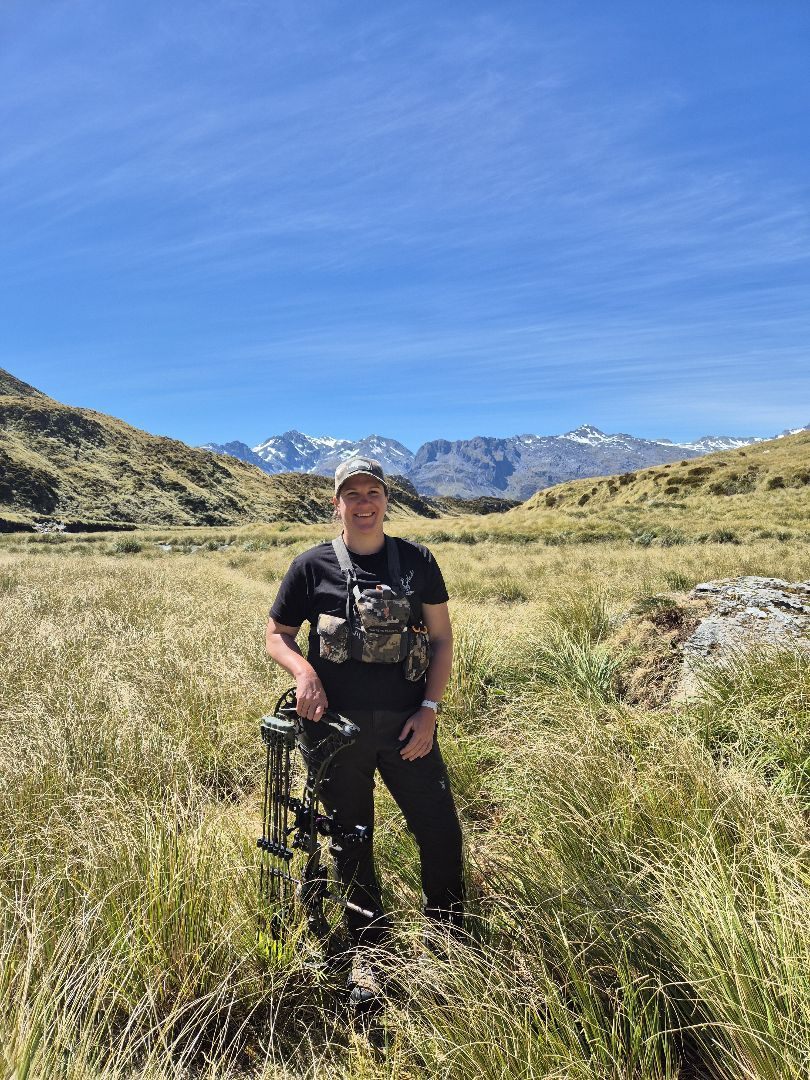 Kate standing in NZ tussock with her bow, snow-capped mountains behind her