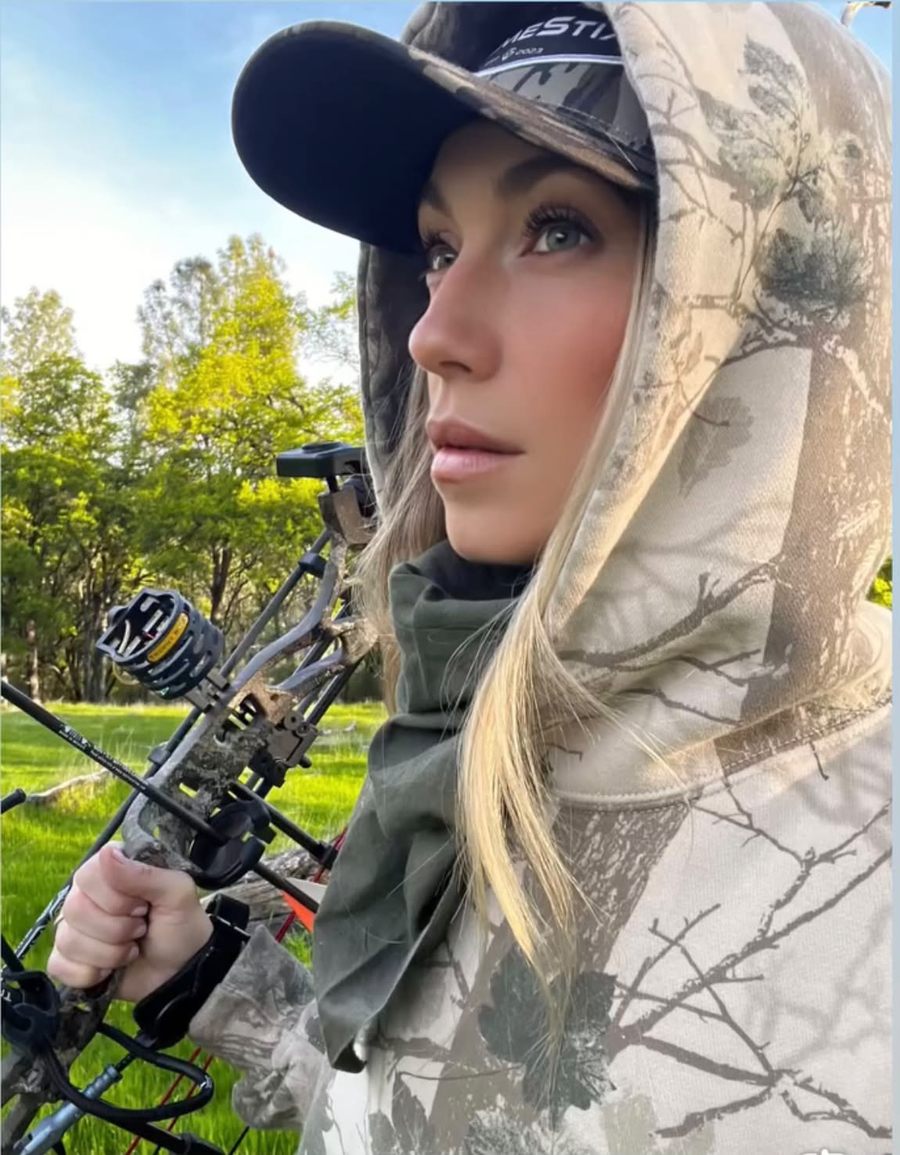 Staysha in a camo hoodie and cap holding her bow in a green meadow