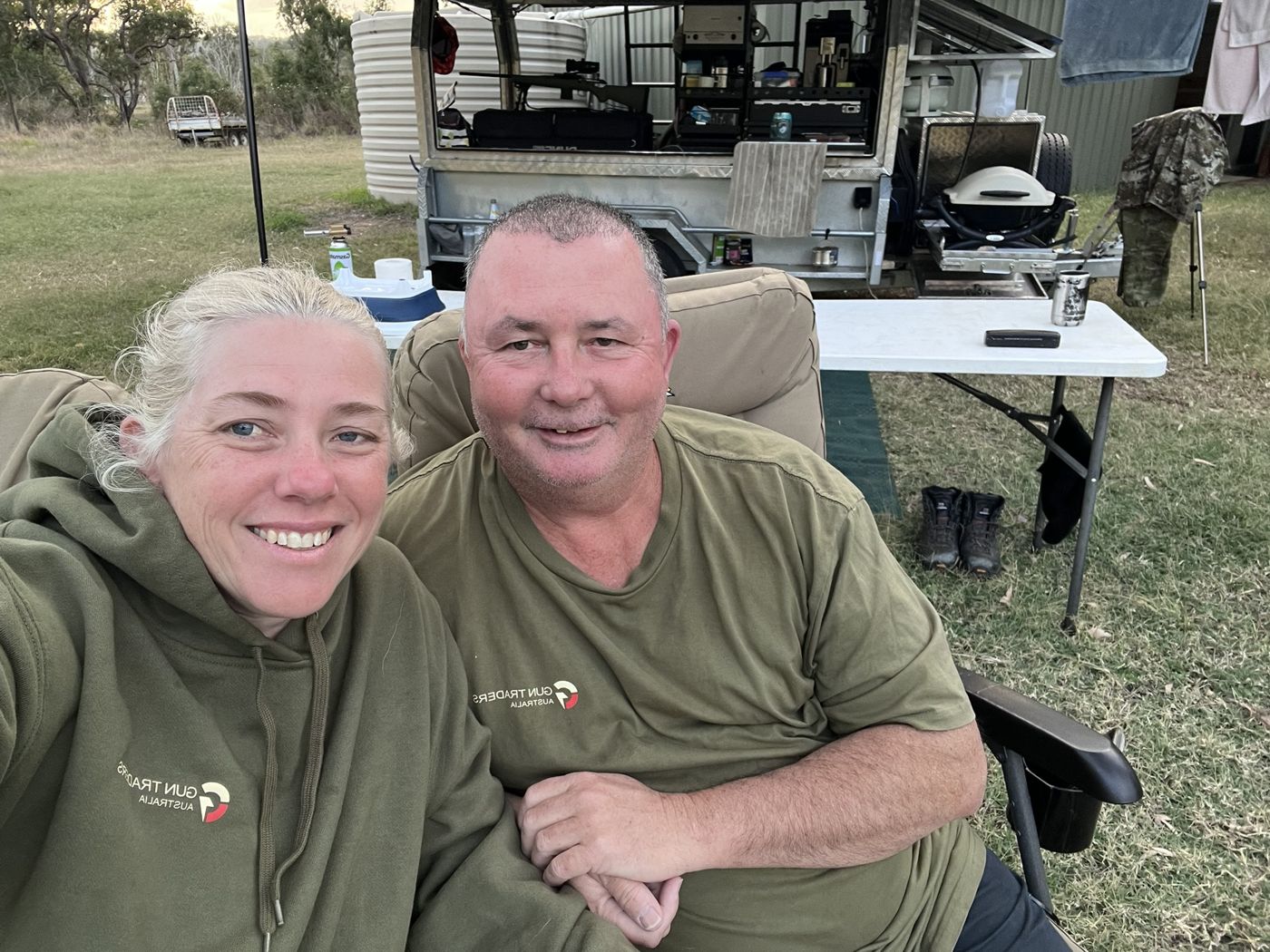 Carly and Glenn in Gun Traders Australia shirts sitting in camp chairs in front of their camper trailer