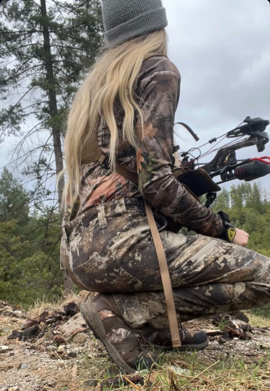 Staysha kneeling in camo on a rocky ridge with her bow, looking out over pine forest