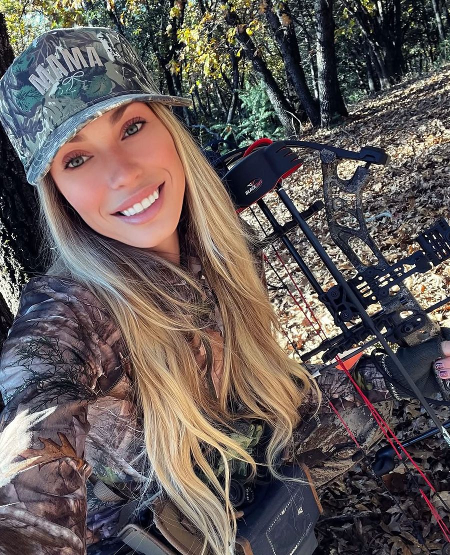 Staysha smiling in a camo MAMA cap with her bow among autumn leaves