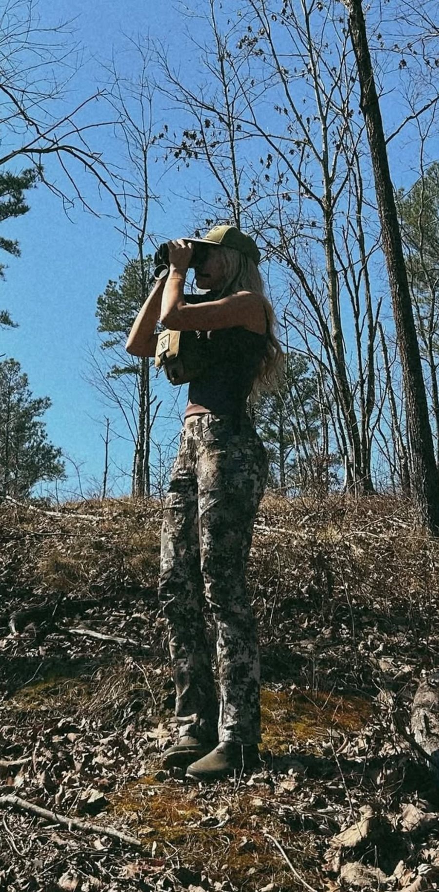 Staysha glassing through binoculars on a wooded hillside under a blue sky