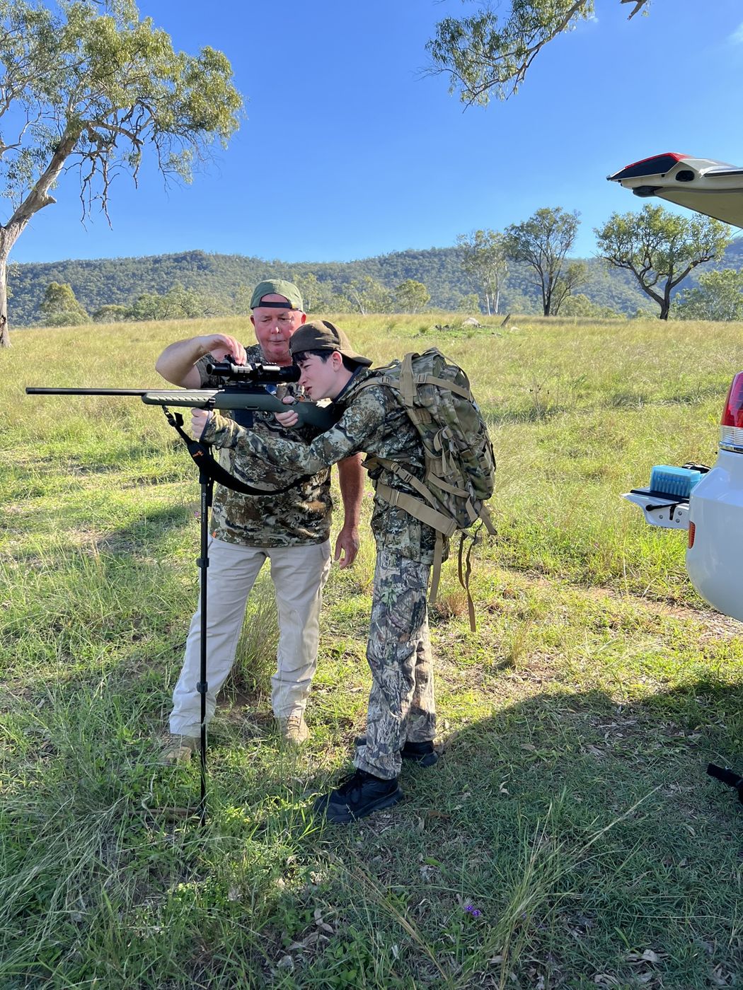 Glenn helping Ethan look through a rifle scope on shooting sticks in an open paddock