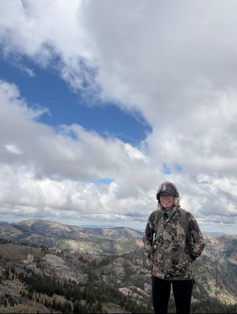 Staysha in a camo jacket and pack on a high ridge with mountains and clouds behind