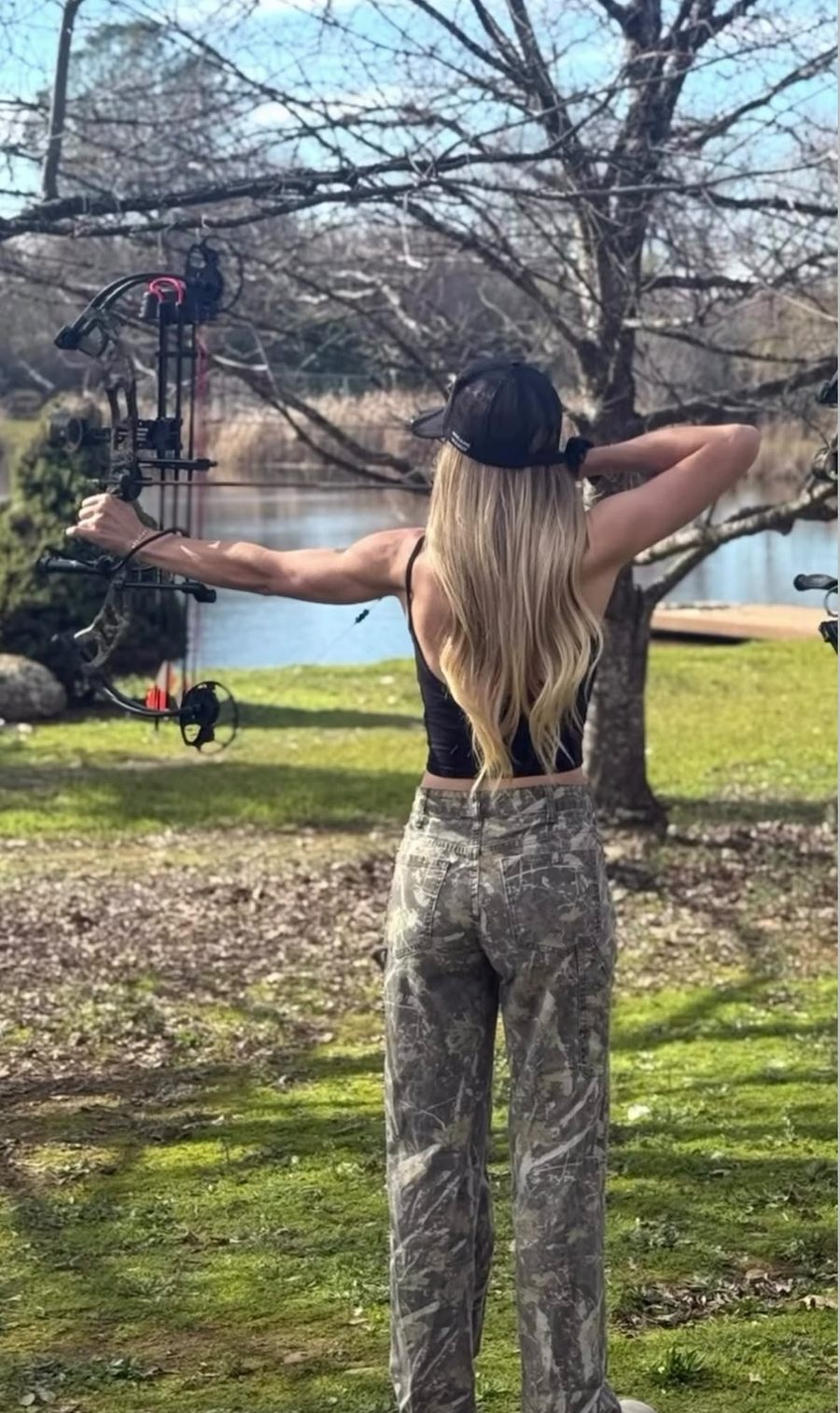 Staysha at full draw with her compound bow on a lawn beside a lake