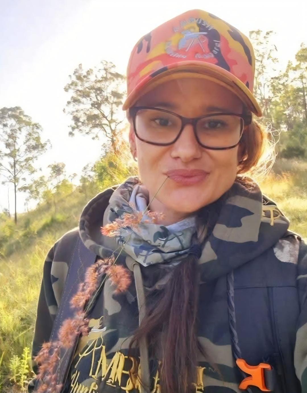 Gaby in her Ambush Hunting Apparel cap and camo hoodie, sunlit grass behind her